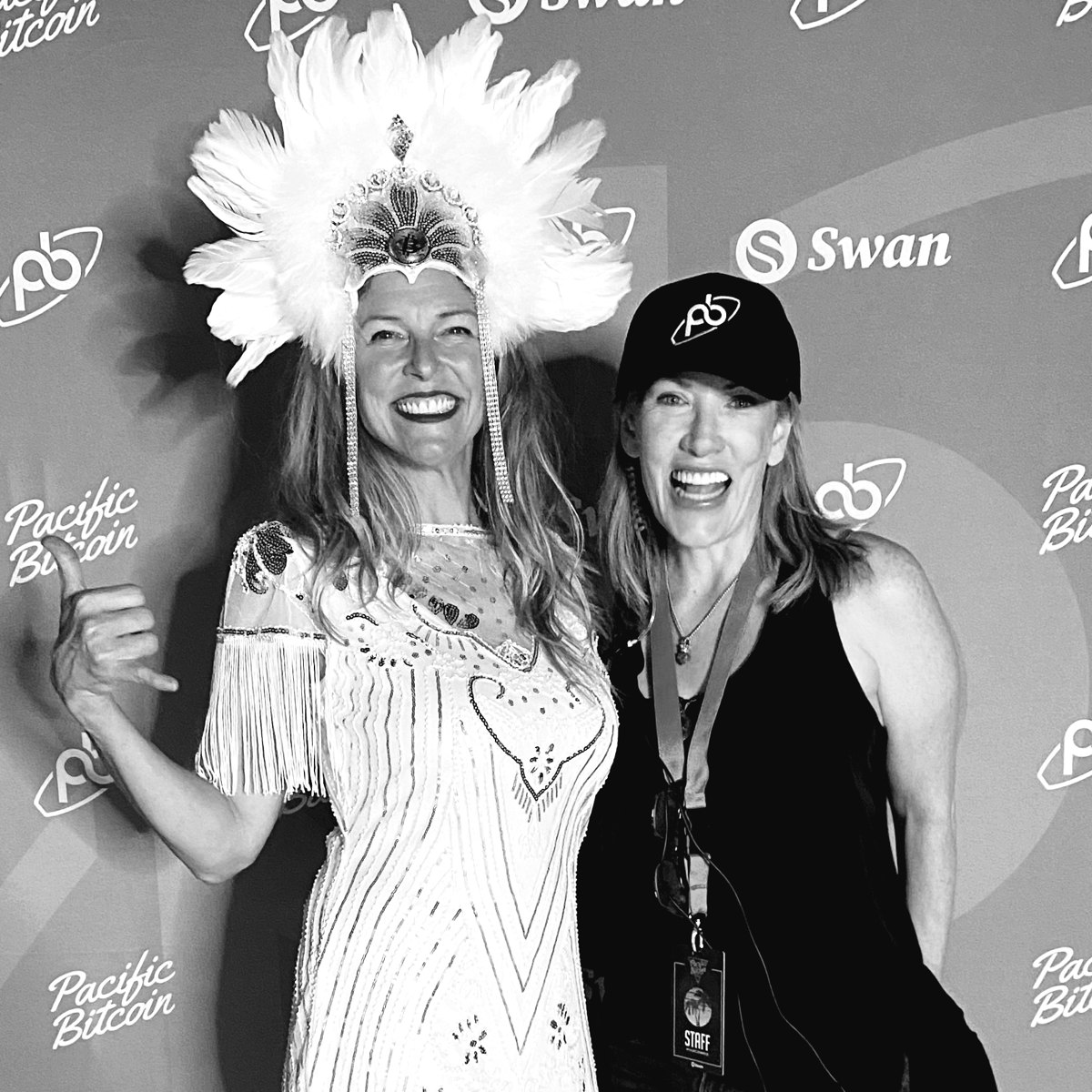 Kristin Thompson in a Pacific Bitcoin cap and staff lanyard, laughing beside a friend in a white feathered headdress.