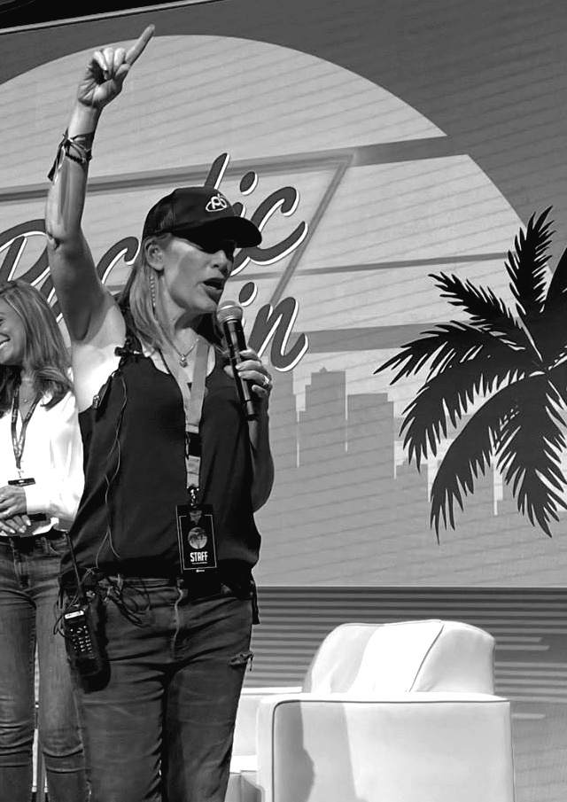 Kristin Thompson on stage at Pacific Bitcoin with a staff badge, radio and microphone, arm raised, running the room.