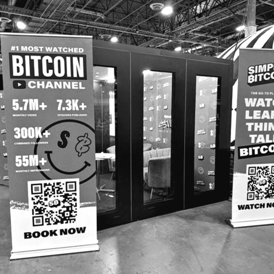 A Simply Bitcoin podcast booth with glass studio rooms and Book Now banners at a conference.